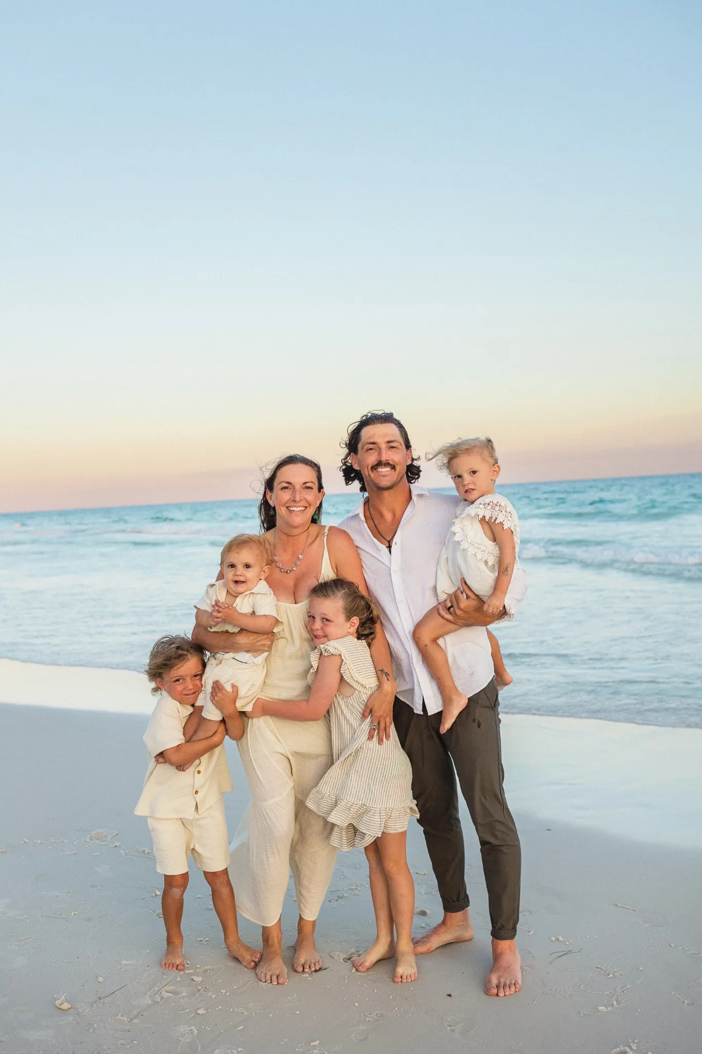 Hailey and Jeremy Aliff standing together on a white sand beach at dusk with their four children gathered around them
