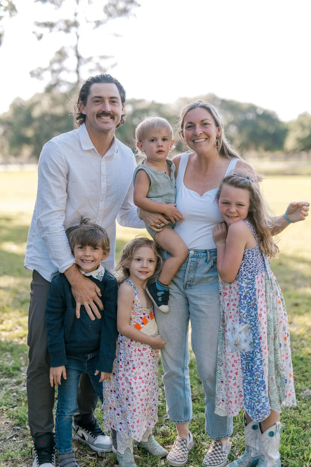 The Aliff family of six standing together in a sunlit park, Hailey holding Jude on her hip