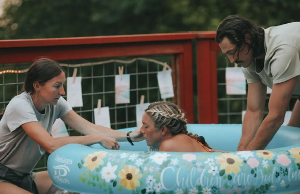 A mother laboring in a garden birth pool, her midwife holding her hand at the rim while her partner leans in beside her