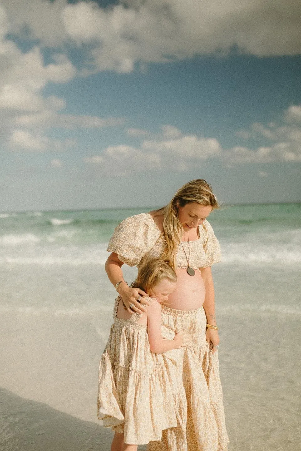 Hailey standing at the water’s edge in a floral dress, her daughter Emerson pressing her face against her pregnant belly