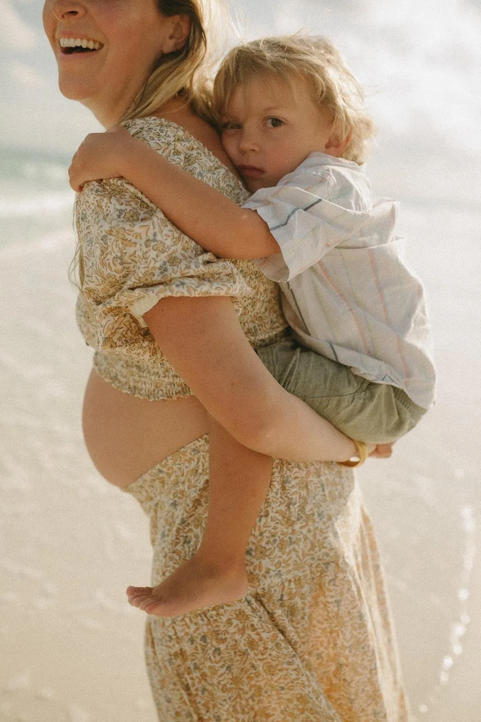 Hailey laughing as she carries her son Koa on her hip at the beach, pregnant with her fourth