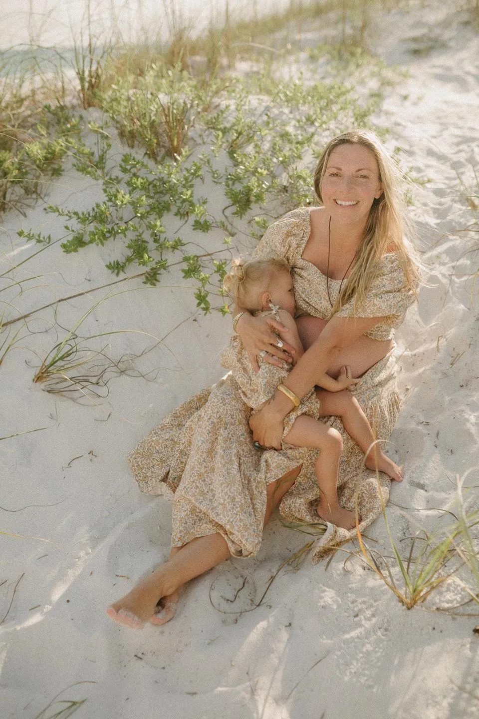 Hailey sitting in the dune grass and sand, holding her daughter Harvest in her lap